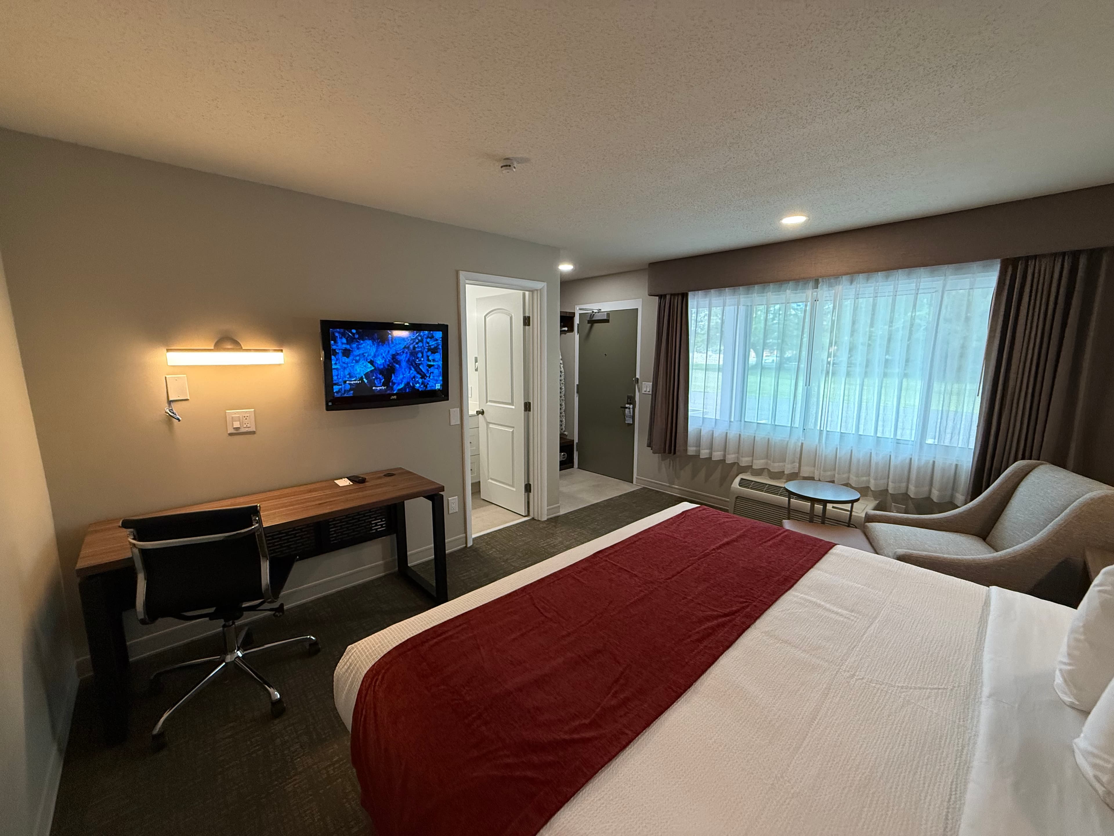 A cozy hotel room featuring a king-sized bed with a red comforter, a wall-mounted TV over a desk, and a sitting area by the window.