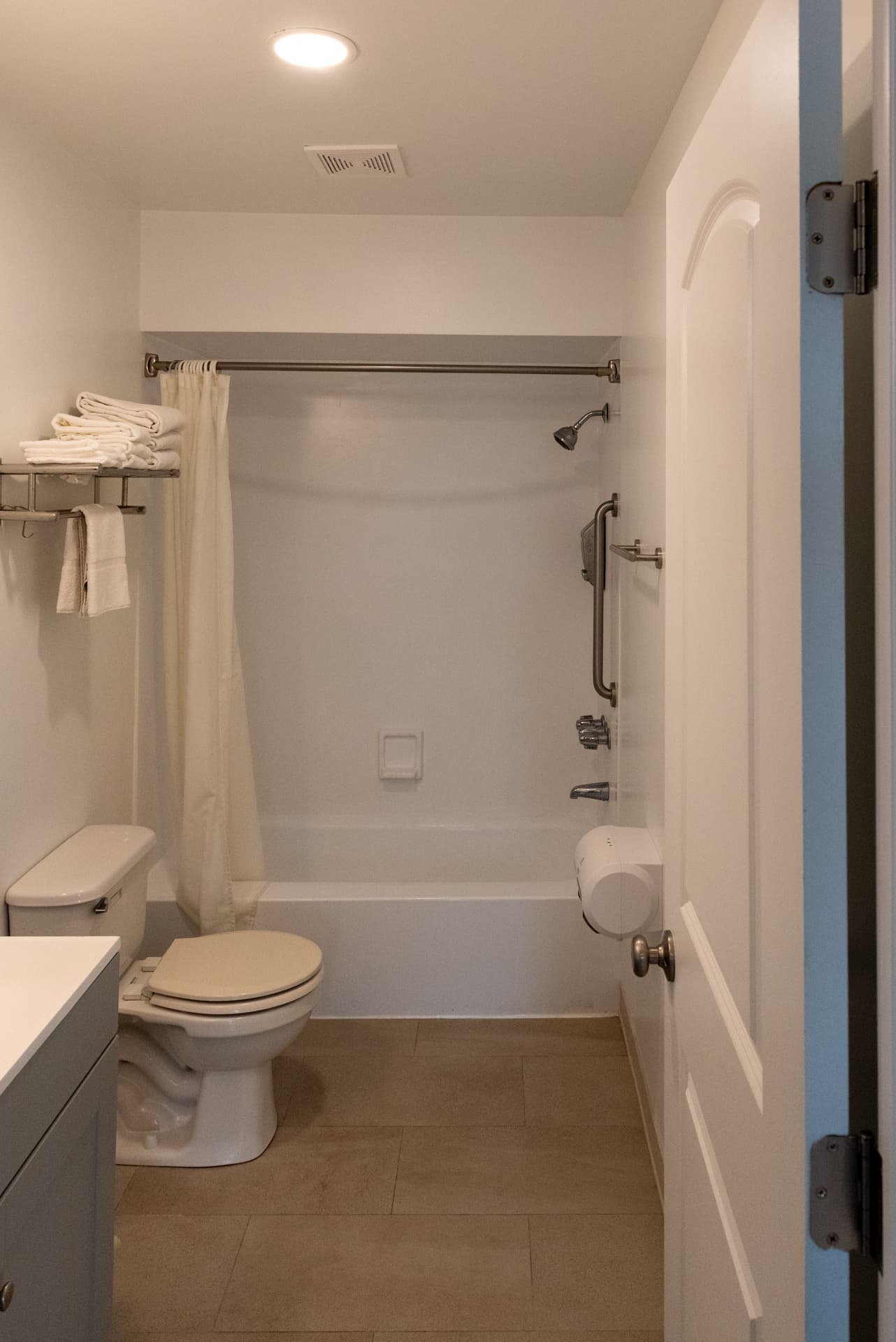 A well-lit hotel bathroom featuring a toilet, a bathtub with a shower, grab bars, and towels on a rack.