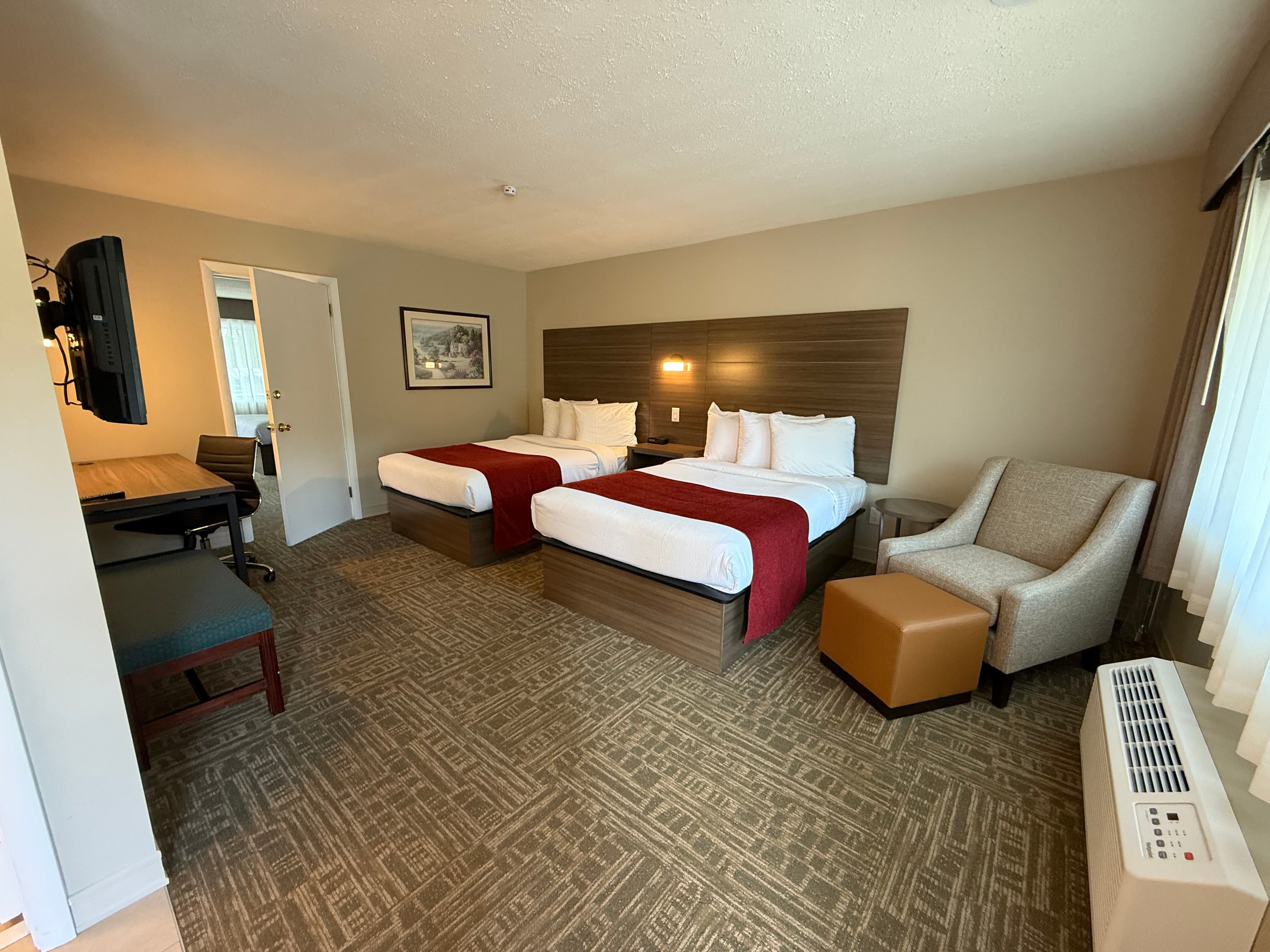 A clean hotel room featuring two queen-sized beds with white and red linens, a desk with a chair, and a cozy armchair with an ottoman.