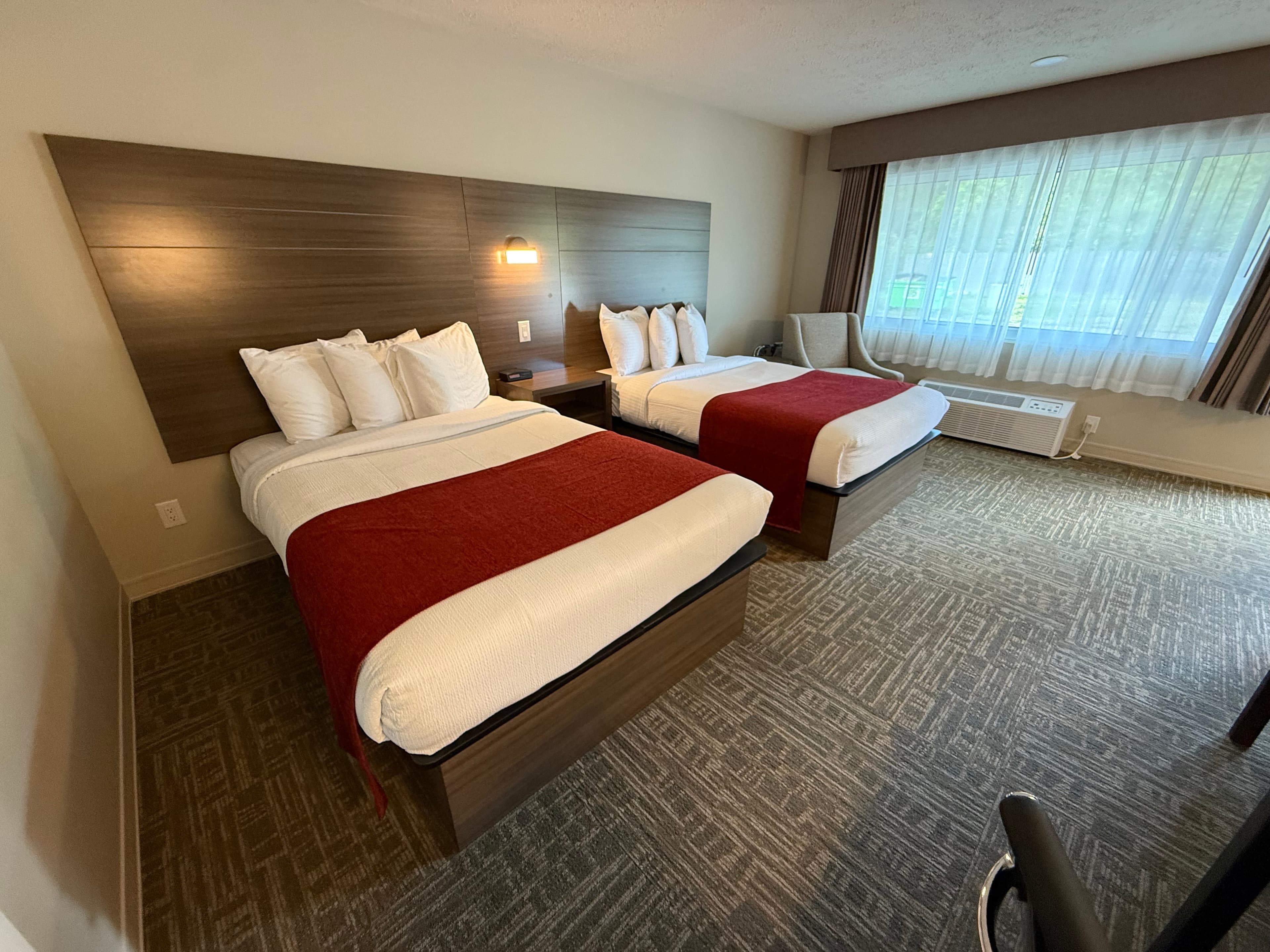 A bright hotel room with two queen-sized beds featuring a minimalist design, a large window with drapes, and a neutral patterned carpet.