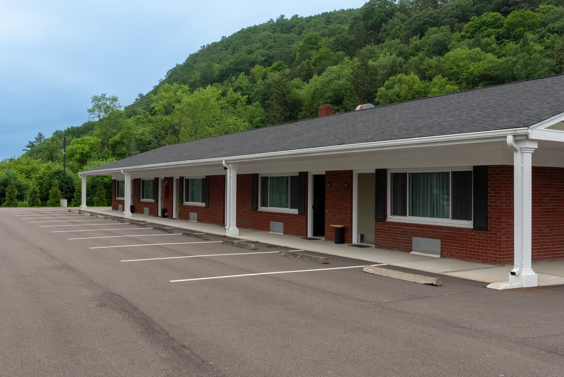 A classic, one-story motel building with a red brick exterior, a covered walkway, and a parking lot. It is set against a lush, green hillside.