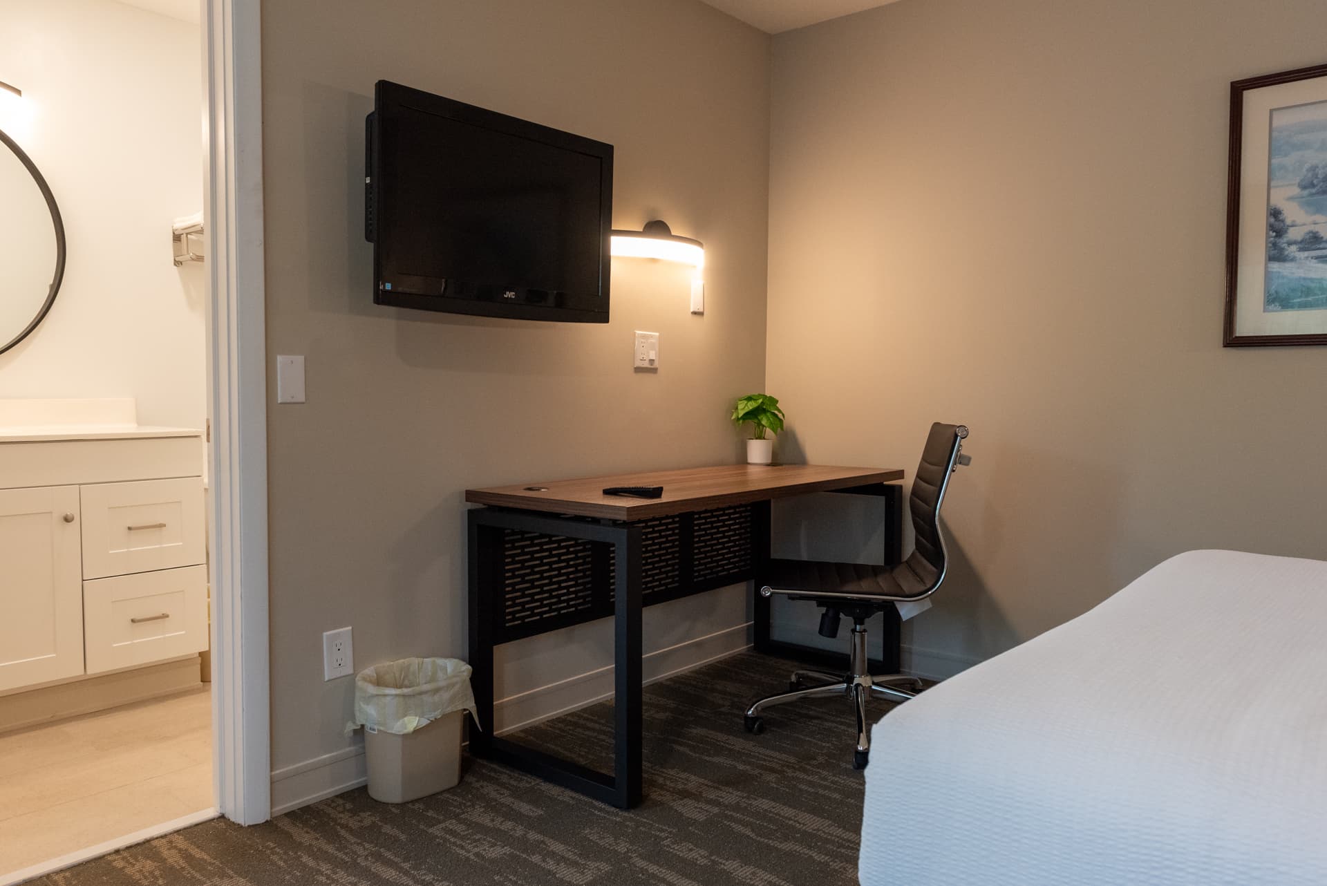 A cozy hotel workspace with a desk, office chair, and a wall-mounted TV. A doorway to a bathroom with a modern vanity is visible.