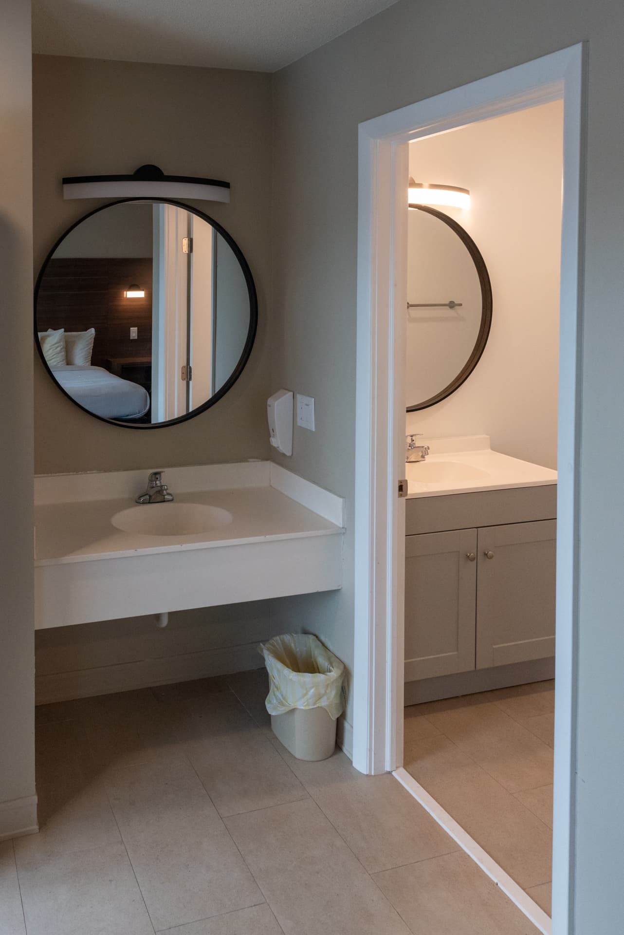 A split hotel bathroom with a separate sink and vanity area, featuring a large mirror and modern lighting. The main bathroom is visible through the doorway.