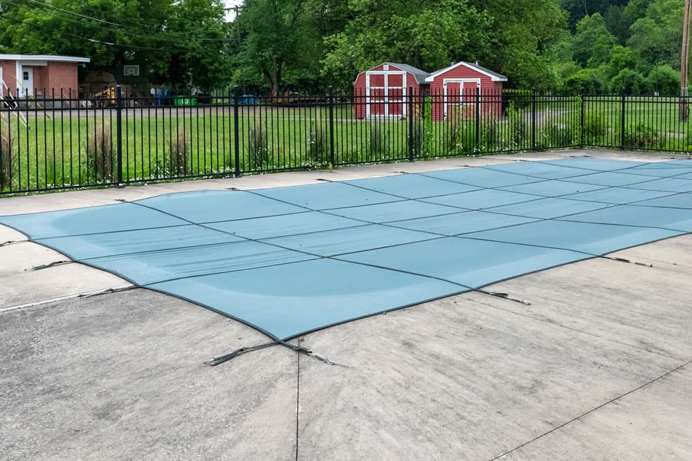 A blue pool cover is spread over a concrete deck, bordered by a black fence and grassy area.